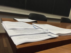 a photo of several piles of class hand outs laid on top of each other on top of a wooden desk with a black chalkboard in the background and grey office swivel chair tucked behind the desk