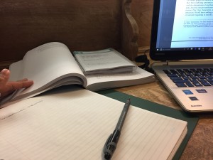 a photo of an open binder with an open textbook slightly on top of it with a day planner opened on top of the textbook sitting beside a laptop on a wooden study desk