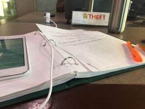 A photo of a white tablet sitting on top of an open green binder filled with notes with a fluorescent orange highlighter at thelower right corner of the open binder. beside the binder at the top right corner is a stainless steel water bottle and beside it a sign that has "Theft" written in bold, red letters acrossit, beside the sign is a plug and from the blug a white cord runs from a charger to the bottomof the tablet.