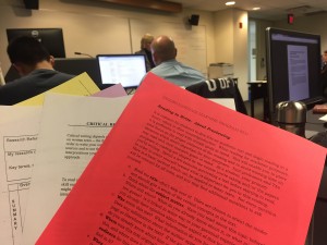 a photo of a stack of hand outs fanned out held in front of a row of students and the instructor wtih a red sheet of paper at the very front