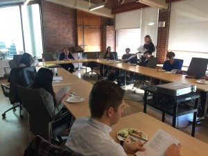 a photo of a seminar class sitting in a board room, each student is sitting at long pressed wood tables and the wall at the end of the room is red brick and copper