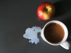 a photo of a mug of coffee sitting on a grey counter taken from above, beside the coffee sits a red and yellow apple and also a splash of spilt milk.