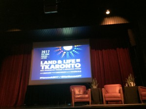 a photo of an empty stage with a few red armchairs and a large projector screen that has the 2017 Hancock Lecture title projected on to it