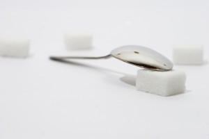 an image of a coffee spoon lying a top a sugar cube with a sugar cube in the distant background, everything sitting on a white surface