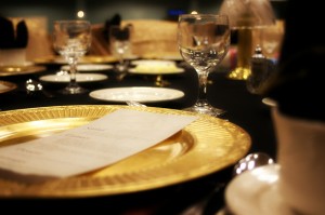 a photo of a dinner place setting with a gold plate and a white dinner roll plate and a wine glass on a table covered with a black table cloth