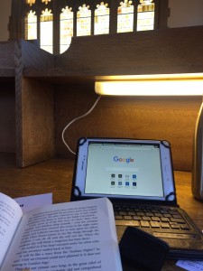 a photo of a work space on a light wooden desk where in the bottom left corner is a top portion of a novel, behind that there is a tablet on the Google homepage in a black case with a keyboard, and besie it is a part of a stainless steel water bottle all backdropped by a large gothic style yellow window