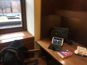 a photo of a wooden work cubicle with a navy bag sitting against the cubicle dividers, infront of the bag is a white table with a black case, lying open with a keyboard, and a purple novel lying beside it, in the bottom left of the picture is a wooden chair with a black wool coat hanging off of it and a black, grey, and purple stiped scrarf hanging as well, and in the tope left corner is a corner of a window that looks out on to a garden and a brick building.