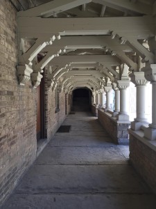 a photo of the UC quad out door hall that leads to the junior common room, there are white ornate pillars along the right side of the hall and a brick wall on the left, above the hall or more white ornate beams that repeat until you get to a wooden door at the end of the hall