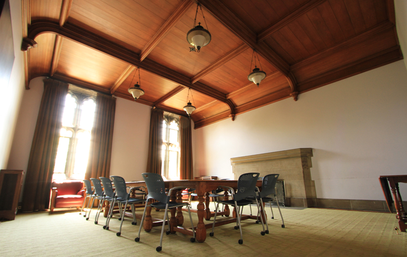 Interior of the Hart House Committees Room. In the foreground: a large, wooden table sits in the center with chairs surrounding it. In the background, the two windows of the room are streaming sunlight in.