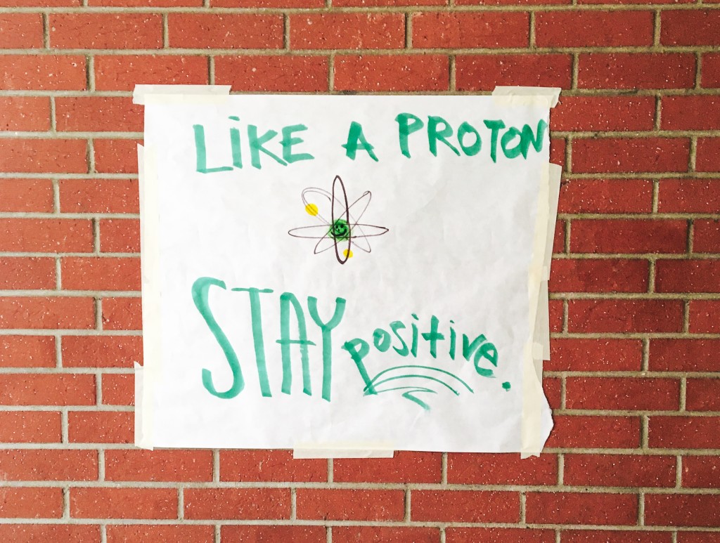 A poster on a brick wall that reads "Like a Proton, Stay Positive!"