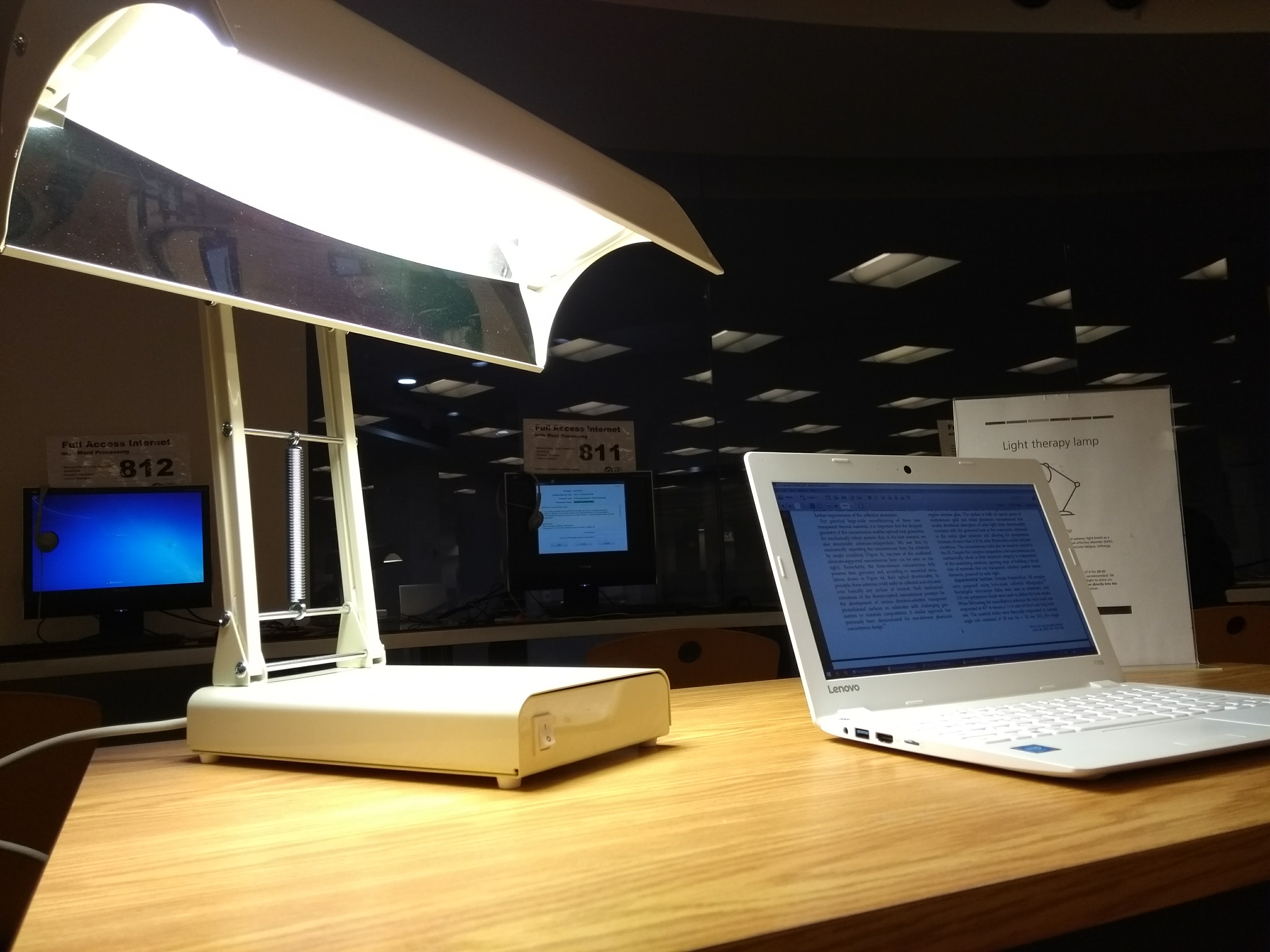 A bright concave-shaped lamp called the Seasonal Affect Disorder (SAD) Lamp on a library desk with a laptop beside it.