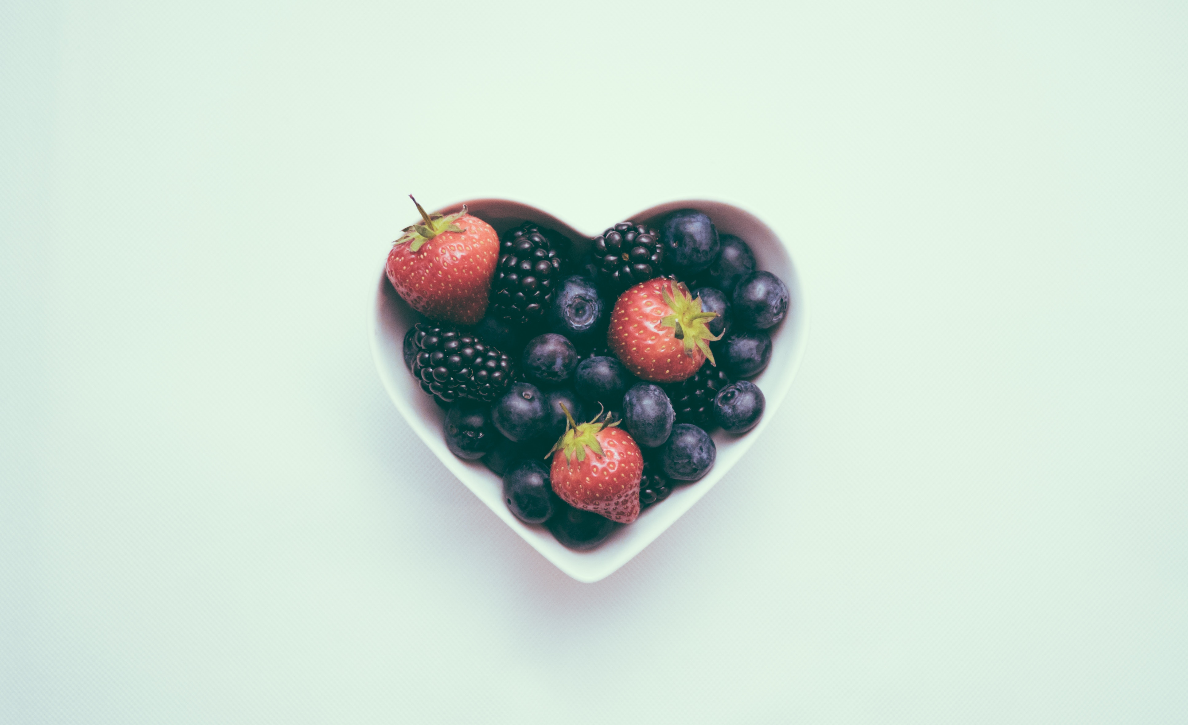 A heart-shaped bowl on a pastel blue background holds some strawberries and blueberries. 