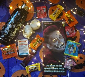On a purple and orange pumpkin tablecloth, there's a waterbottle, two cloves of garlic, a temporary scary smile face tattoo, a tube of fake blood, a card that lists emergency resources around the community, and a bunch of candy. This is all surrounded by a string of lights.