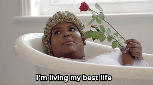 person in a bathtub with a bonnet on, holding a rose. Caption reads "I'm living my best life"