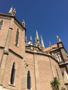 Photograph: large Spanish colonial style church, light brick color, some gothic influences, blue sky background, top of palm tree in lower right hand corner
