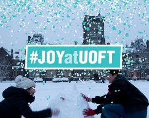An image of two people building a snowman on front campus, with the blue words #JoyAtUOfT.