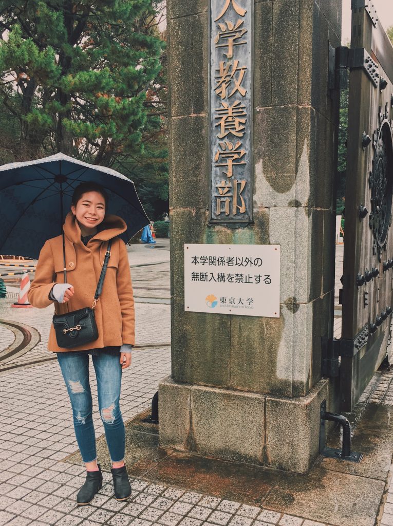 Emi, holding an umbrella, standing in front of the University of Tokyo entrance gate.
