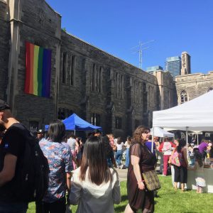 A crowd of people standing near Hart House, with a rainbow flag hung on the building. 