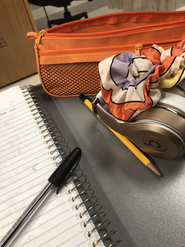 A notebook with a pen lying over it is set down next to an orange pencil case, a pencil, a set of headphones, and a scrunchie.