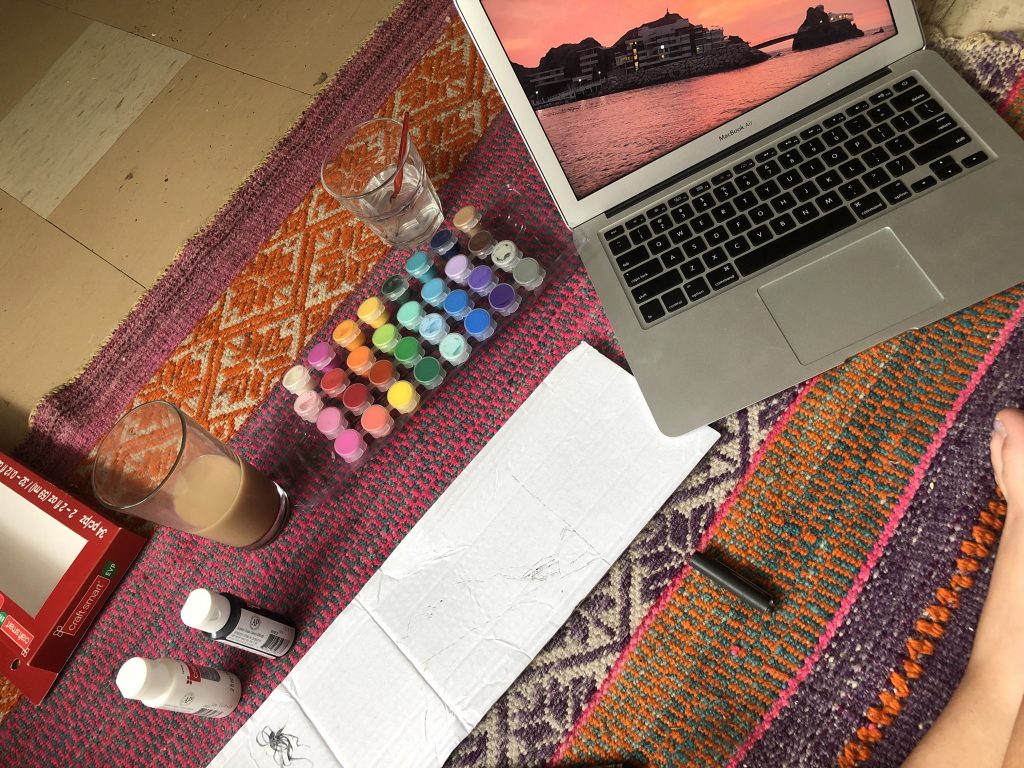 On a colourful carpet acrylic paint is setup next to a laptop with the image of a sunset open on it.