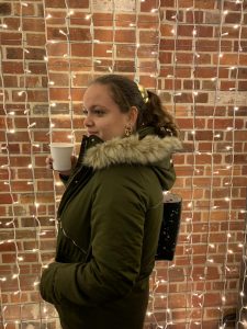 Francesca looking to the side with a cup of hot chocolate in her hands. Behind her there's a brick wall covered by fairy lights.