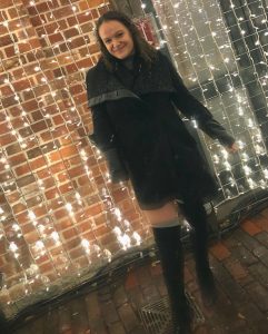 Francesca posing for the camera in front of fairy lights. She's wearing a black coat and black knee-high boots.
