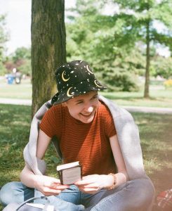 Girl grins in a park while holding a book. She is wearing a hat with the night sky patterned on it.