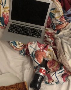 An open laptop on a bed, next to it a bottle of Coke and a slice of Pizza