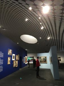 Museum patrons look at paintings hung from blue walls, beneath a domed ceiling with black and white patterns on it. The focal point is a circular skylight.