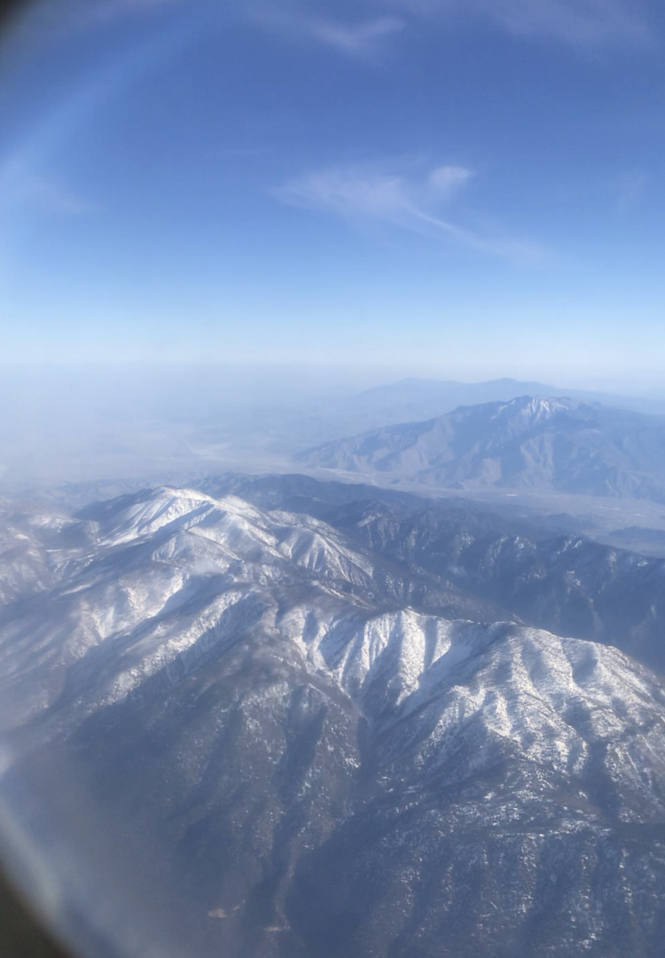 Picture of mountains taken through an Airplane window. There is snow on the tops of the mountains and a vast landscape