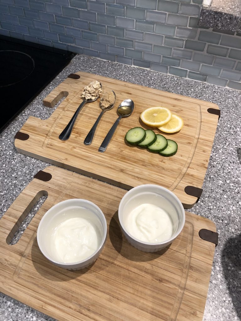 Chopping board with lemon slices, cucumber, spoons filled with honey, oats, and sugar. Two bowls of yogurt.
