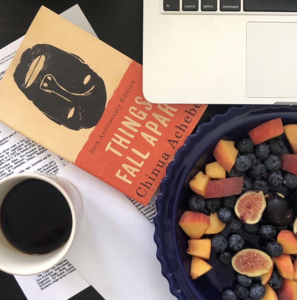 An aerial photo of a desk of someone that's been studying. In the frame, a black coffee, a plate of fruit (peaches, blueberries and figs) and a book, Things Fall Apart by Chinua Achebe.
