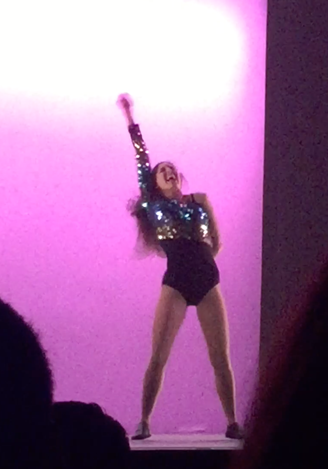 Sammi with her hand in the air during dance performance, pink background, sparkly top