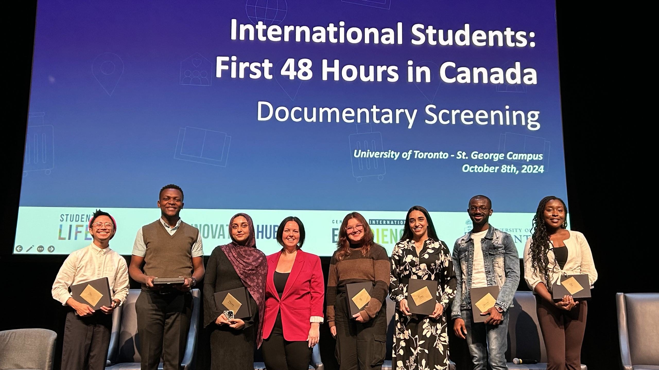 A group photo from the "International Students: First 48 Hours in Canada" documentary screening at the University of Toronto, featuring eight attendees standing on stage, smiling, and holding plaques.