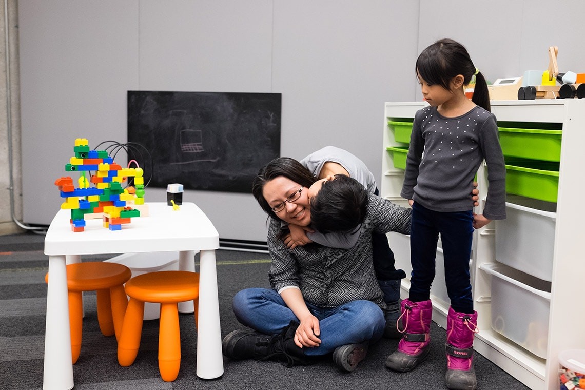 An student parent sitting on the floor, cross-legged with a child hugging them. Another child standing beside them, in the Robarts Family Study Space. A black chalkboard, child-sized furniture and toys are in the background.
