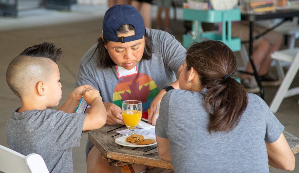 A family of three sitting around a table doing a Pride activity and enjoying snacks.