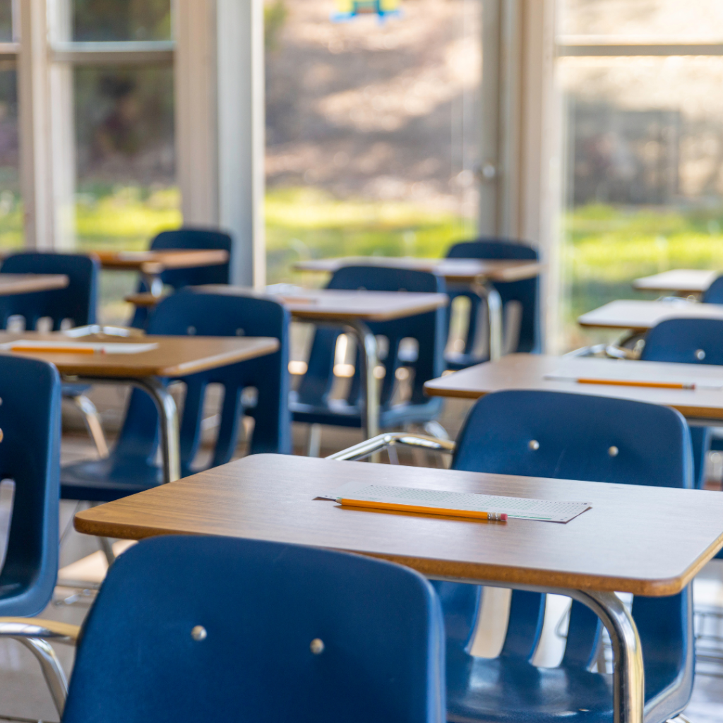 Image of wooden school desks with blue chairs. There is a scantron and pencil on each seat. There are 3 rows of seats shown. The back of the classroom there are windows. It is daytime.