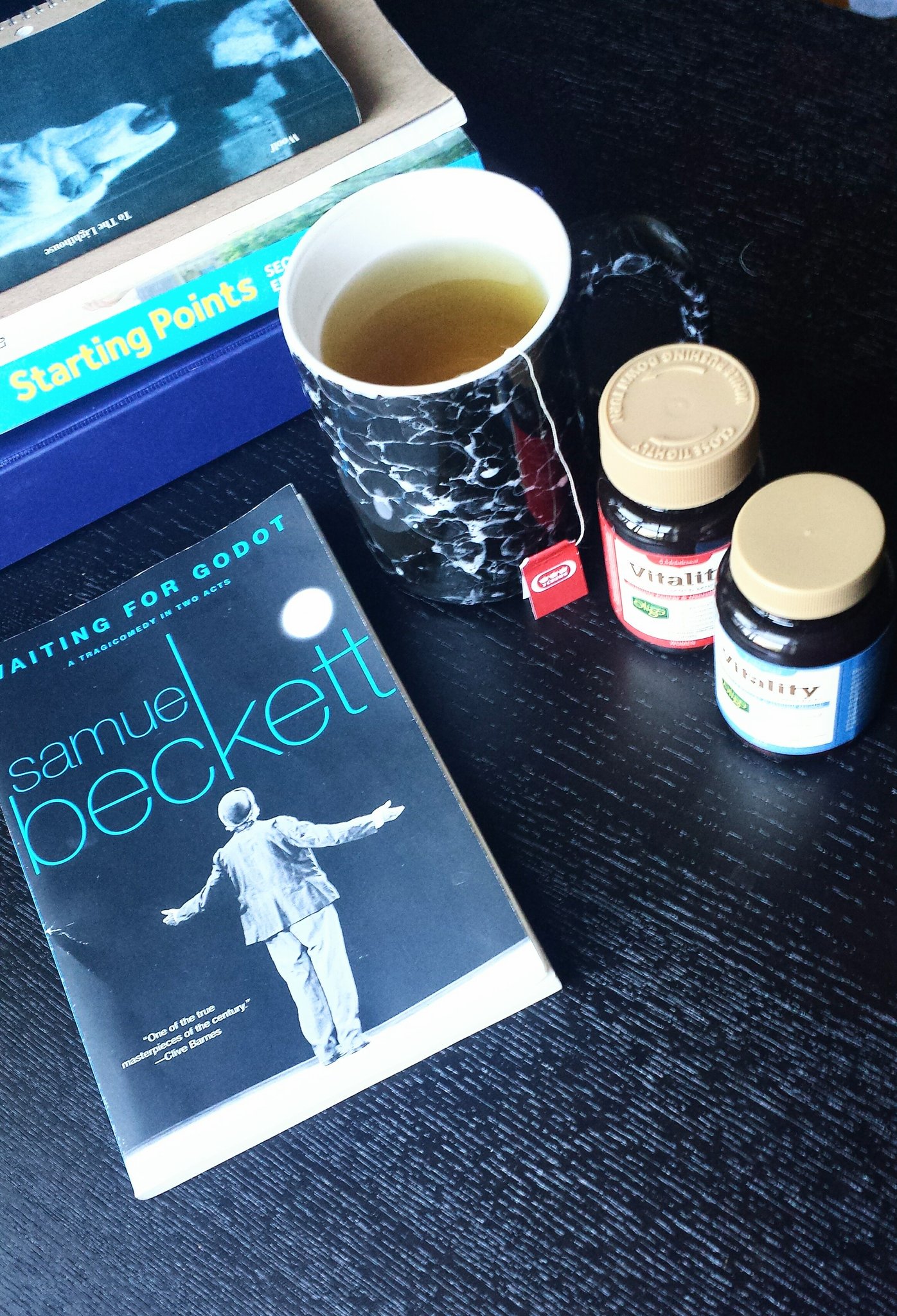 An image of a mug of tea, two containers of multivitamins and the playbook "Waiting for Godot" assorted on a black coffee table.