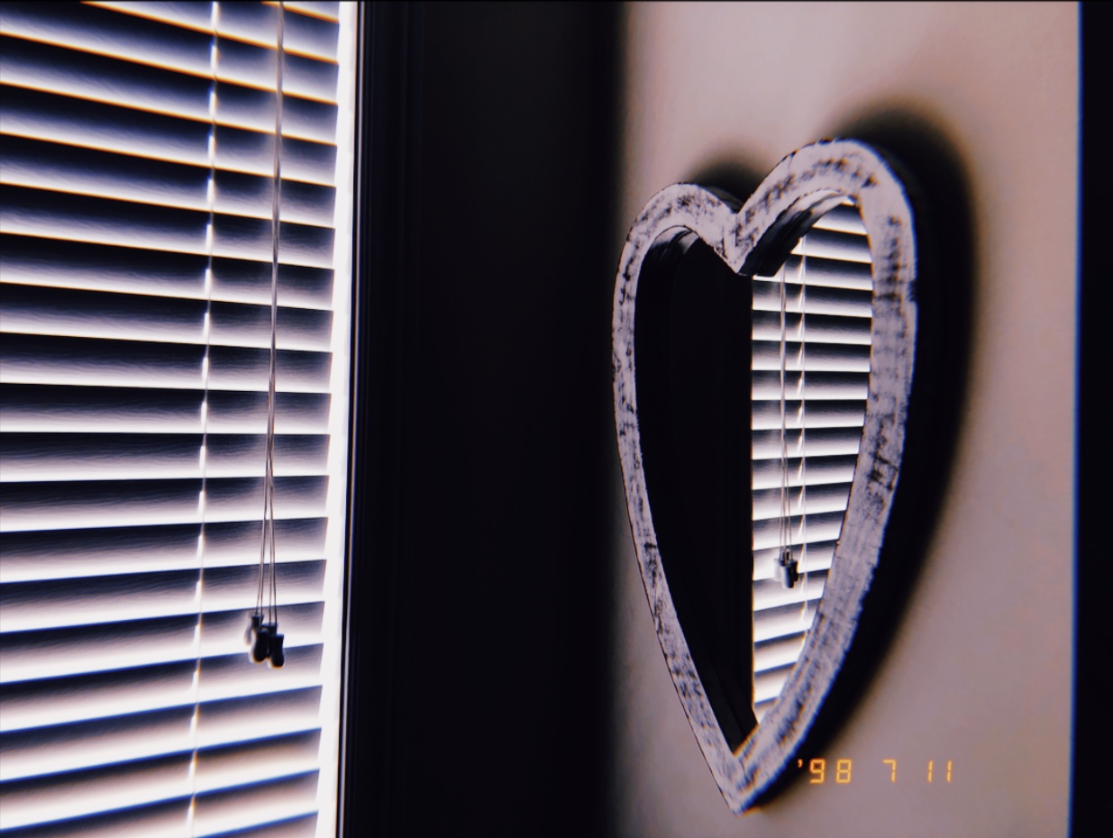 image of a heart shaped mirror handing on a wall by the corner. The reflection shows closed blinds that let the soft glow of the morning light through.
