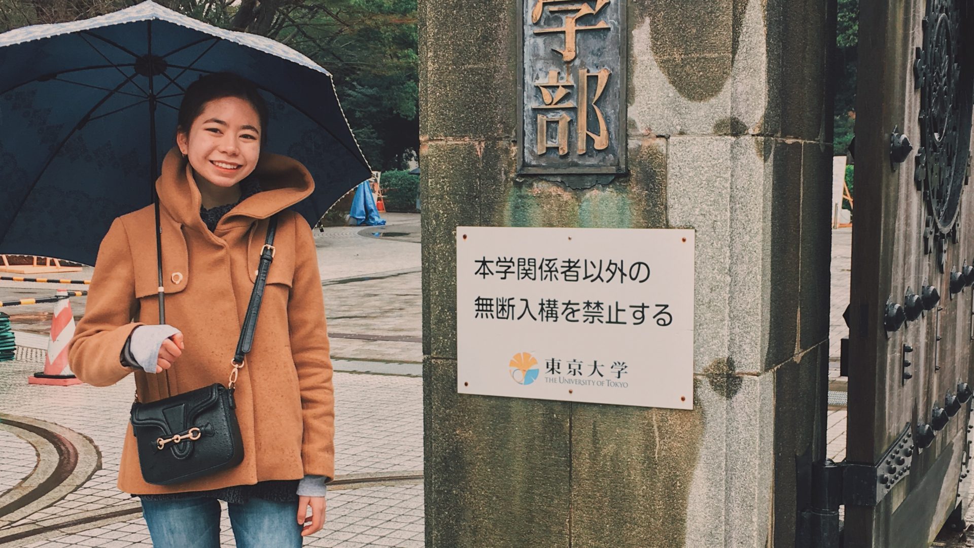 Emi, holding an umbrella, standing in front of the University of Tokyo entrance gate.