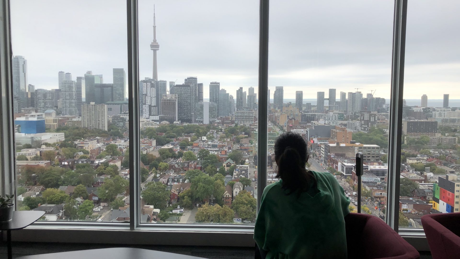 A girl sits by the window of a high-rise building and you can see the skyline in the background.