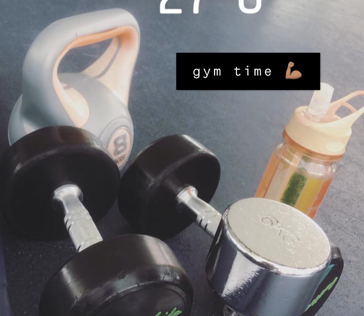 A couple of dumbells and a water bottle set on the floor. There's a water stamp that reads 27 degrees celcius. There's also text with a black background that reads "gym time".