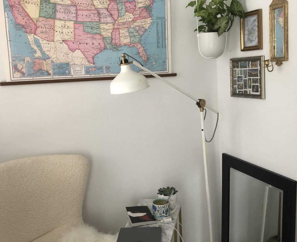 A cranny in a room, with a map of the United States on the wall, a large arm chair in the corner, and a standing lamp hovering over a marble table.