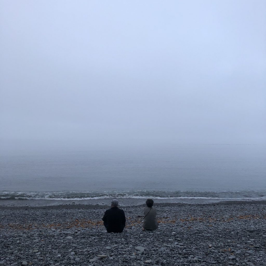 Two people, with their backs turned, sitting on a foggy beach.