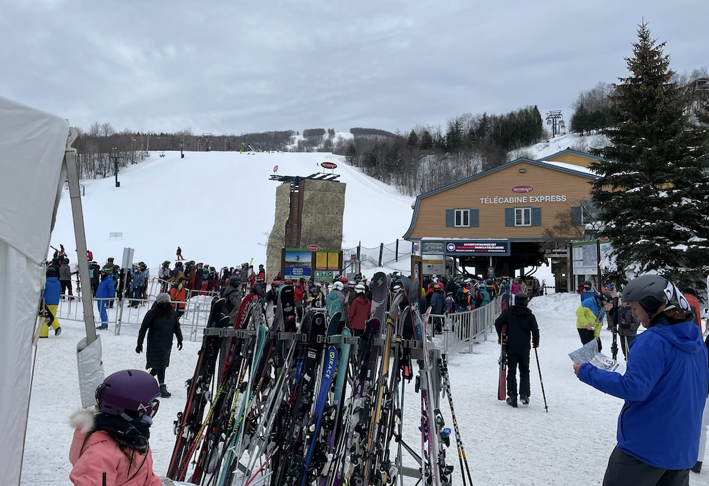 many skis and poles lined up at a rack at the top of a busy ski mountain with people