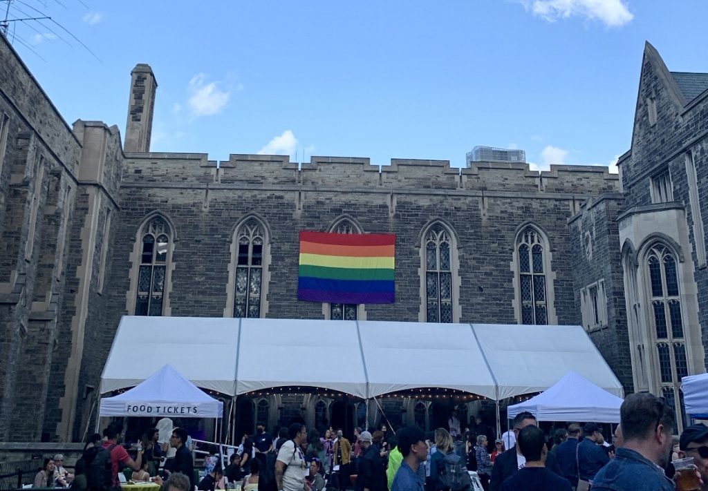 Crowd of people gathered outdoors, a white large tent behind them and a Pride flag hanging in the background.
