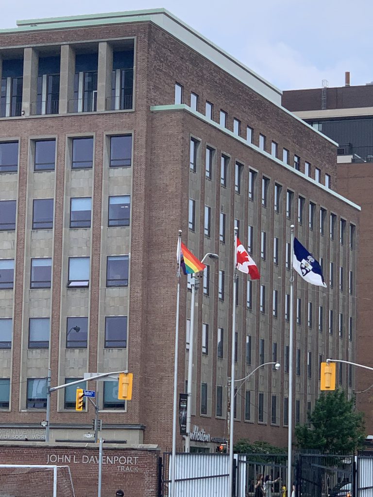 Three waving raised flags: the Progress Pride Flag, Canada Flag, and U of T Flag.