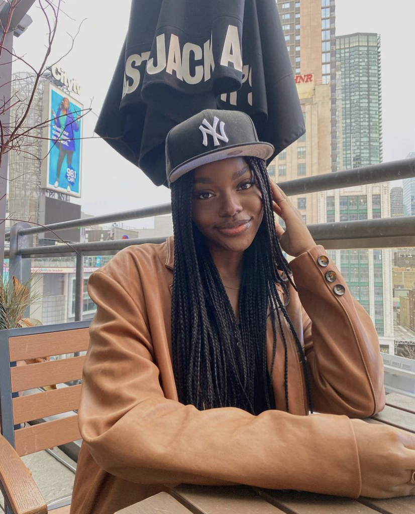 Crystal wearing a New York Yankees baseball hat and a leather jacket, at a patio dining table outdoors. 